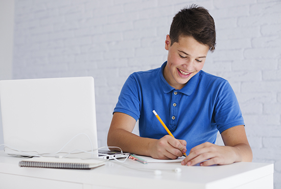 Un hombre joven camiseta azul sentado en un escritorio con un computador ey escribiendo en un cuaderno.