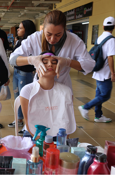 Una persona de pie hace un masaje en la frente y rostro a una estudiante universitaria que está sentada en una silla 