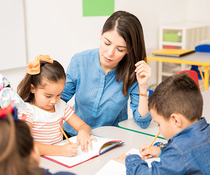 Conmemorando la niñez con experiencias significativas En un salón de clases, profesora acompaña a una niña a hacer su actividad. La infante tiene su cuaderno abierto y está realizando la acción de escribir o dibujar.