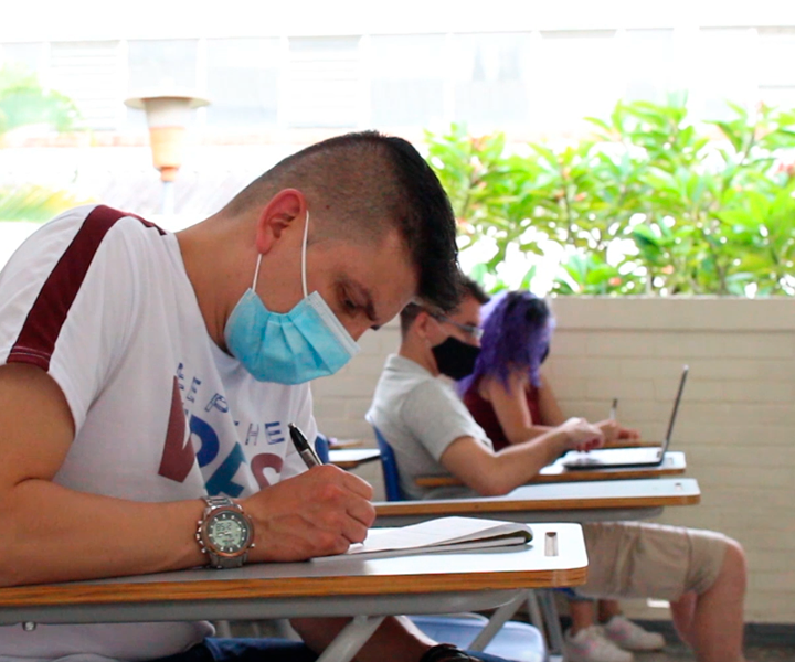 Estudiante usa tapaboca y escribe en silla de universidad. Al fondo se ven dos compañeros suños escuchando la clase. 