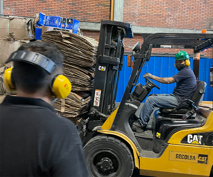 Camión de carga lleva una pila de carton mientras un hombre mira la actividad, usando los elementos de seguridad siguiendo la legislacion en SST