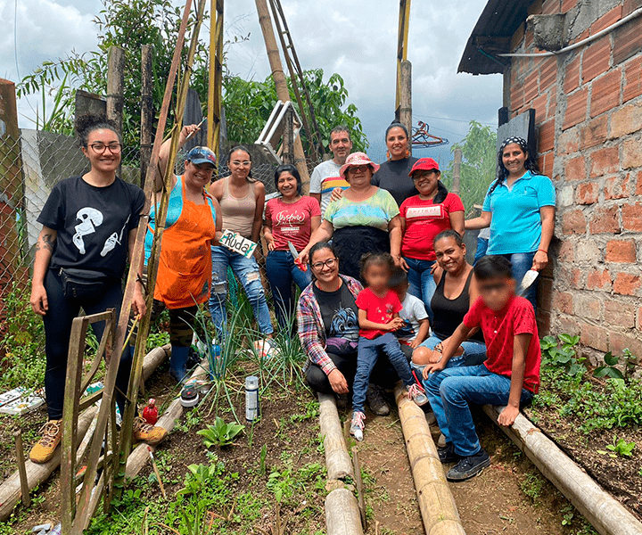 Mujeres en la huerta comunitaria, en la parte inferior de la fotografía se ve el sembrado, planatas verdes, divididas por guaduas. 