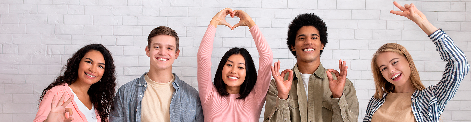 Amigos multirraciales felices adolescentes haciendo gestos con las manos, la gente en equipo casual se divierten y miran la cámara, sobre fondo de pared de ladrillo blanco