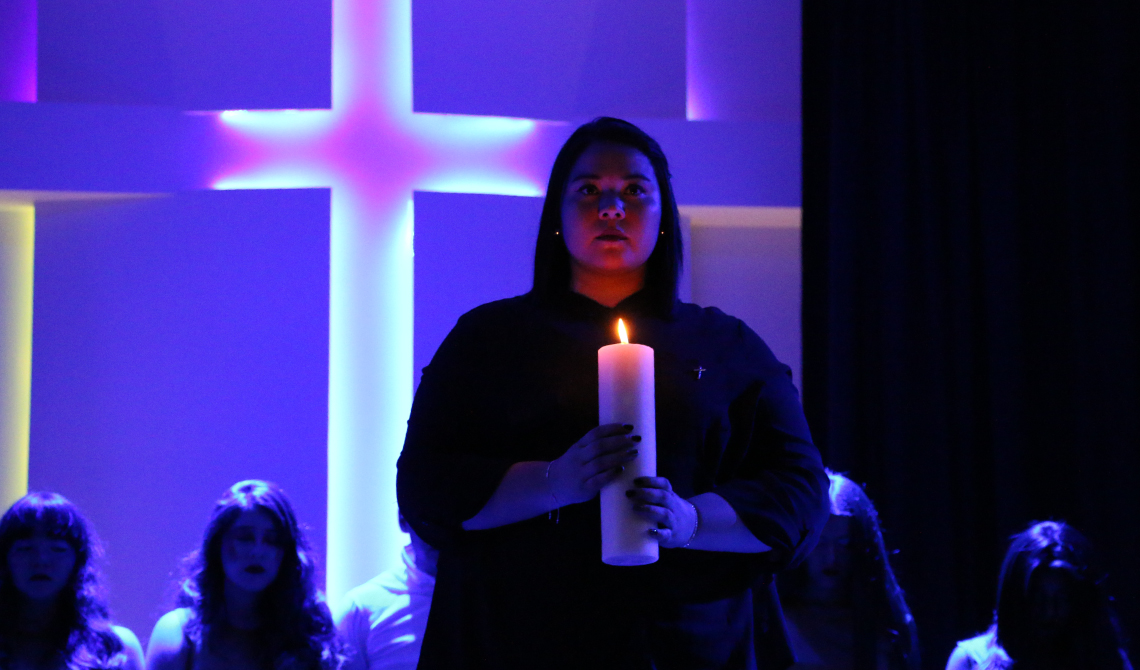 Una mujer sostiene un cirio con sus manos, en el auditorio oscuro. Atrás se observa una cruz de luces. 