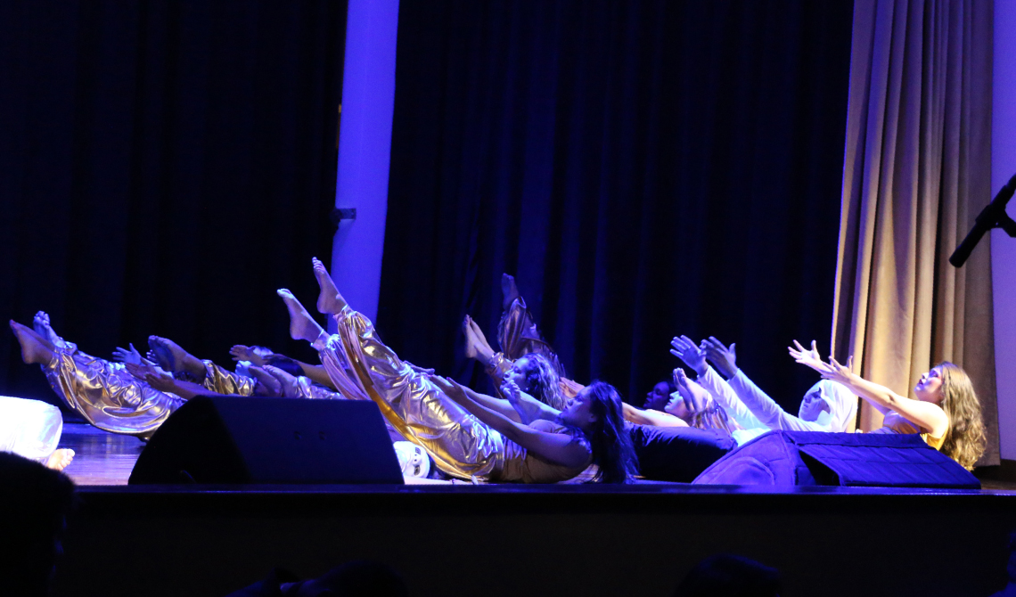 Grupo de danza, acostados en el piso de la tarima con las piernas hacia arriba, realizando su performance. 