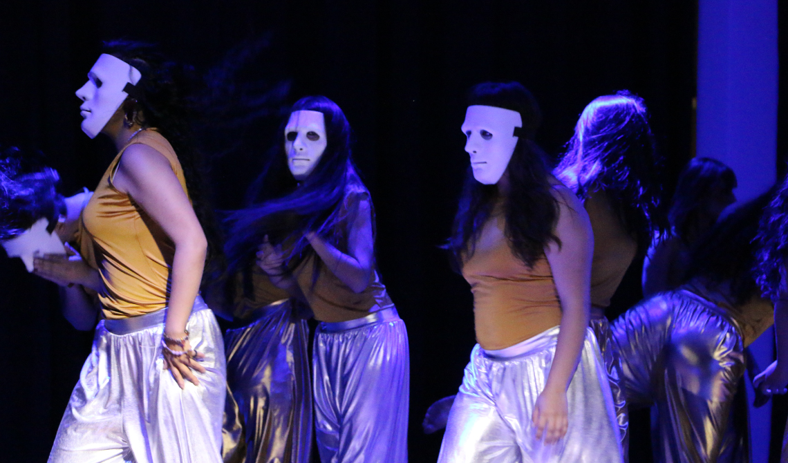 Grupo de danza con máscaras blancas y pantalones brillantes, caminado por la tarima. 