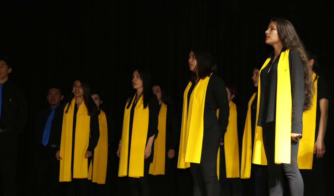 Varias mujeres con prenda amarilla larga que baja desde el cuello y dos hombres con corbata azul cielo. 