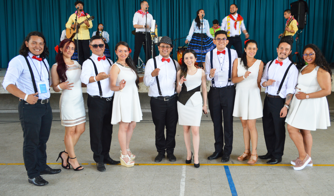 Grupo de baile con trajes elegantes posando para la foto. Más atrás, un grupo de música ensayando. 