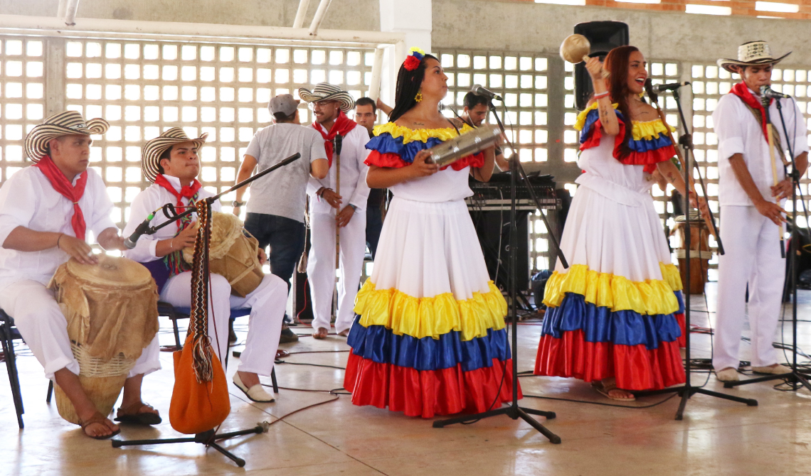 Grupo con trajes de colores de la bandera, cantando y tocando instrumentos de percusión y viento. 