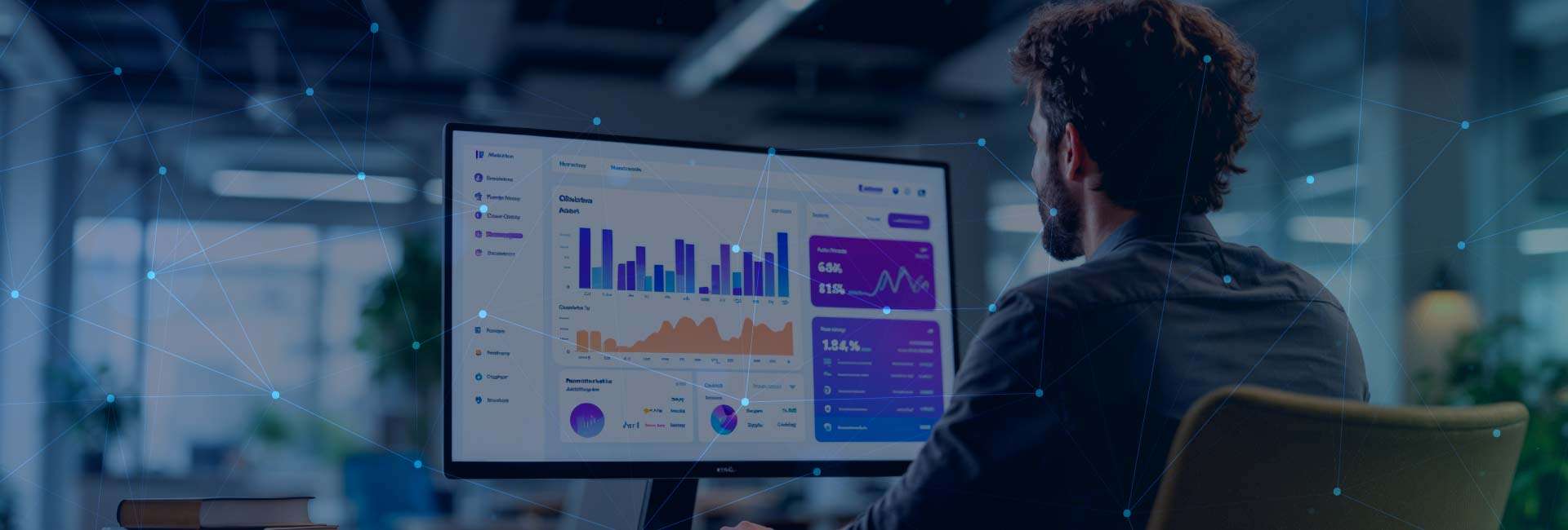 Hombre con camisa oscura sentado en una oficina moderna, trabajando en una computadora con gráficos y estadísticas en la pantalla