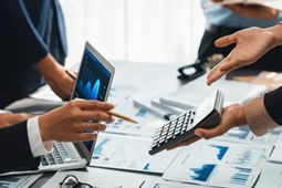 Manos de profesionales revisando gráficos financieros en una laptop y documentos impresos mientras utilizan una calculadora durante una reunión de análisis financiero.