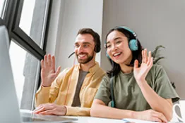 Una mujer y un hombre con audífonos haciendo seña de saludo a un computador