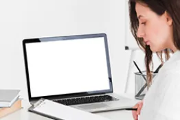 Una mujer sentada en escritorio hay un computador y ella lee un libre.