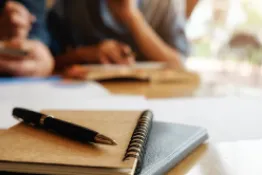 primera plano de cuadernos con lapicero sonre ellos, al fondo se vem 2 personas