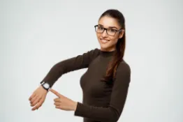 Mujer morena con gafas de cabello largo señalando reloj en su mano derecha