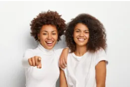 dos jovenes de tez oscura, con cabello crespo, sonrientes y de camisetas blancas, la joven de la izq apunta hacia adelante