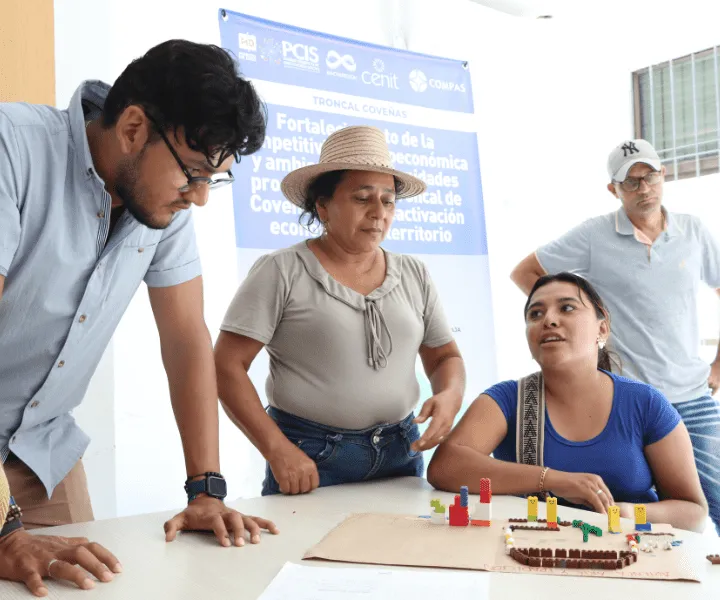 Cuatro personas participan en una actividad grupal en un aula iluminada. Una mujer con sombrero de paja habla mientras los demás la escuchan atentamente. En la mesa hay una maqueta con bloques de colores. Al fondo, un cartel menciona el fortalecimiento económico en la Troncal de Coveñas.