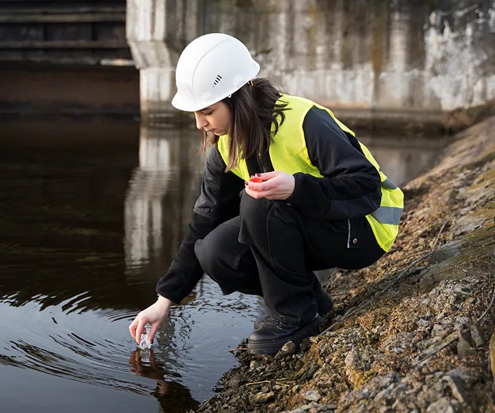 Mujer con casco y chaleco de seguridad recogiendo una muestra de agua en un frasco, para análisis ambiental o de calidad del agua