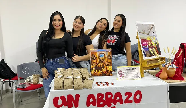 Grupo de estudiantes detrás de una mesa decorada con alimentos y un cartel que dice “Un abrazo de fe”.
