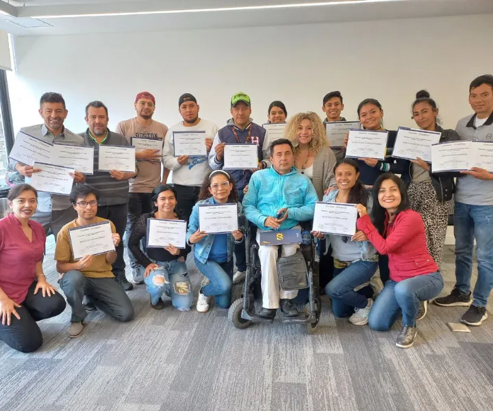 Grupo de personas posando sonrientes en una sala iluminada mientras muestran sus diplomas de graduación o participación.