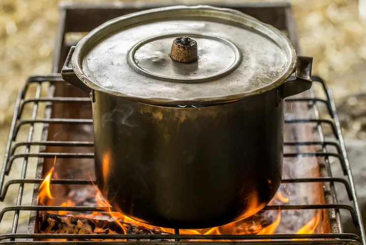 Olla de metal con tapa sobre una parrilla de hierro sobre un fuego generado por una estufa ecológica