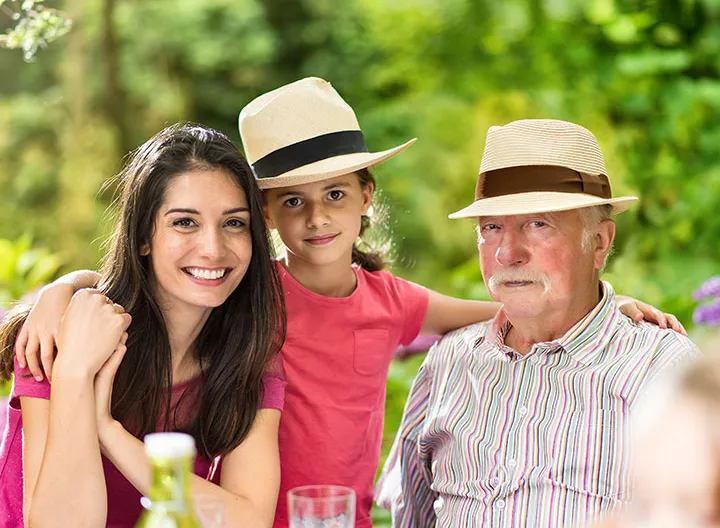 Tres personas reunidas en una foto familiar donde está un señor mayor, una mujer y una niña. Un fondo con naturaleza de color verde