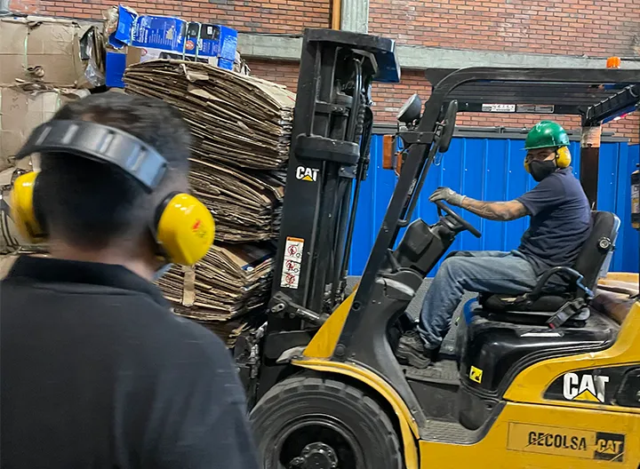 Camión de carga lleva una pila de carton mientras un hombre mira la actividad, usando los elementos de seguridad siguiendo la legislacion en SST