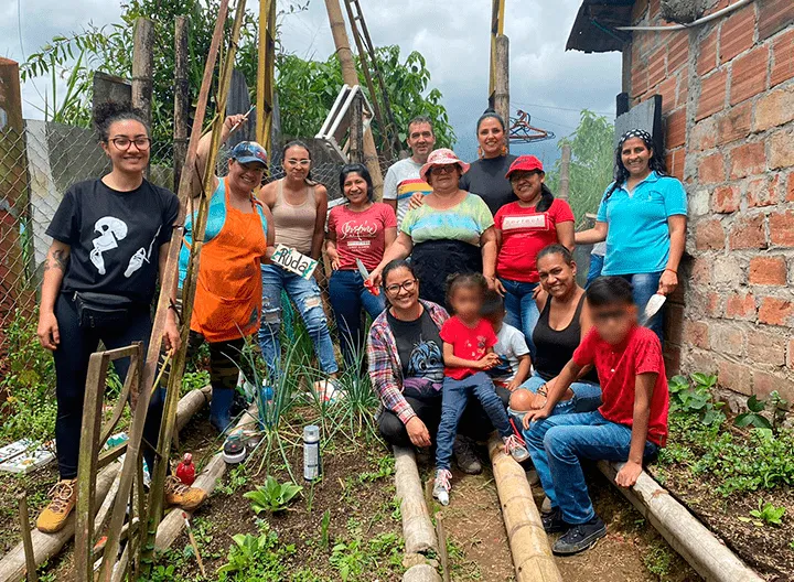Mujeres en la huerta comunitaria, en la parte inferior de la fotografía se ve el sembrado, planatas verdes, divididas por guaduas. 