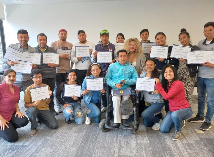 Grupo de personas posando sonrientes en una sala iluminada mientras muestran sus diplomas de graduación o participación.