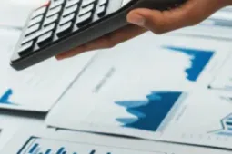 Manos de profesionales revisando gráficos financieros en una laptop y documentos impresos mientras utilizan una calculadora durante una reunión de análisis financiero.