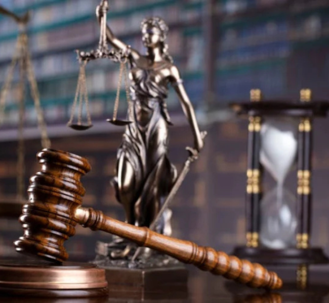 Balanza de la justicia junto a un mazo de juez sobre una mesa, con un reloj de arena y libros de derecho al fondo, representando el concepto de ley, justicia y paso del tiempo en los procesos legales.