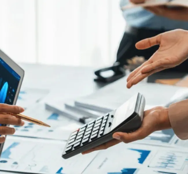 Manos de profesionales revisando gráficos financieros en una laptop y documentos impresos mientras utilizan una calculadora durante una reunión de análisis financiero.